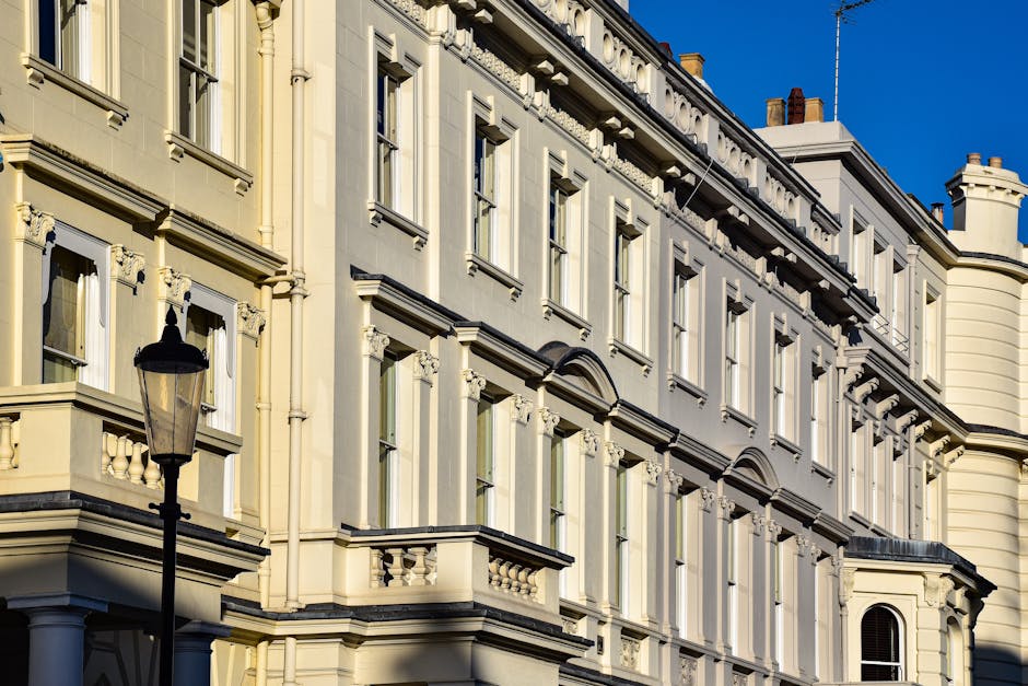 A row of white, Victorian-style terraced houses with detailed cornices, large windows, and small balconies on a city street. Several vehicles, including black, grey, and white cars, are parked in fron